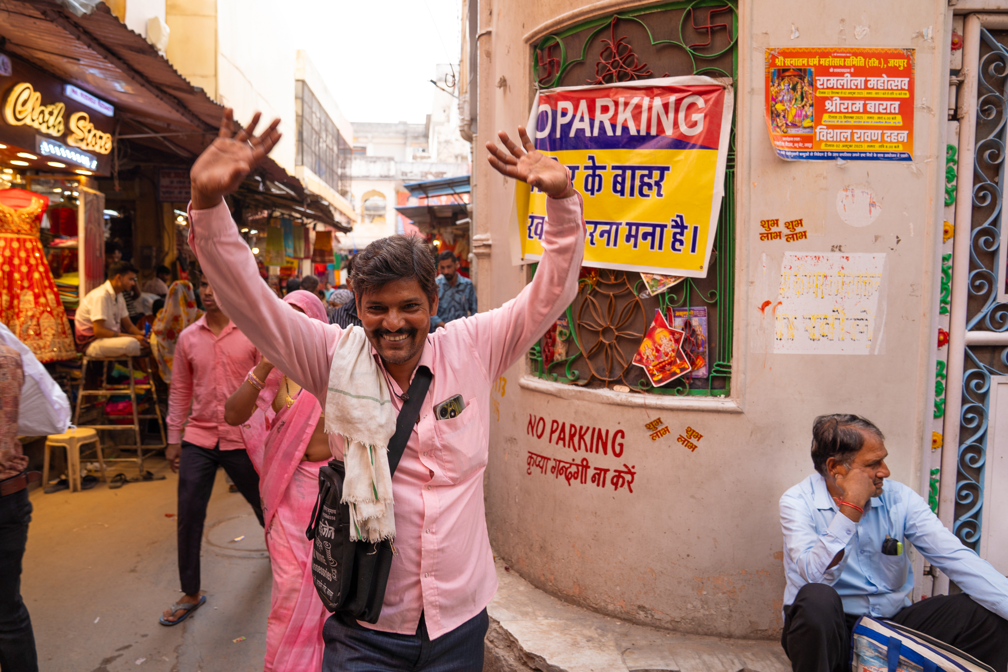 A smiling man in a pink shirt with both arms raised on a busy market street in Jaipur, India