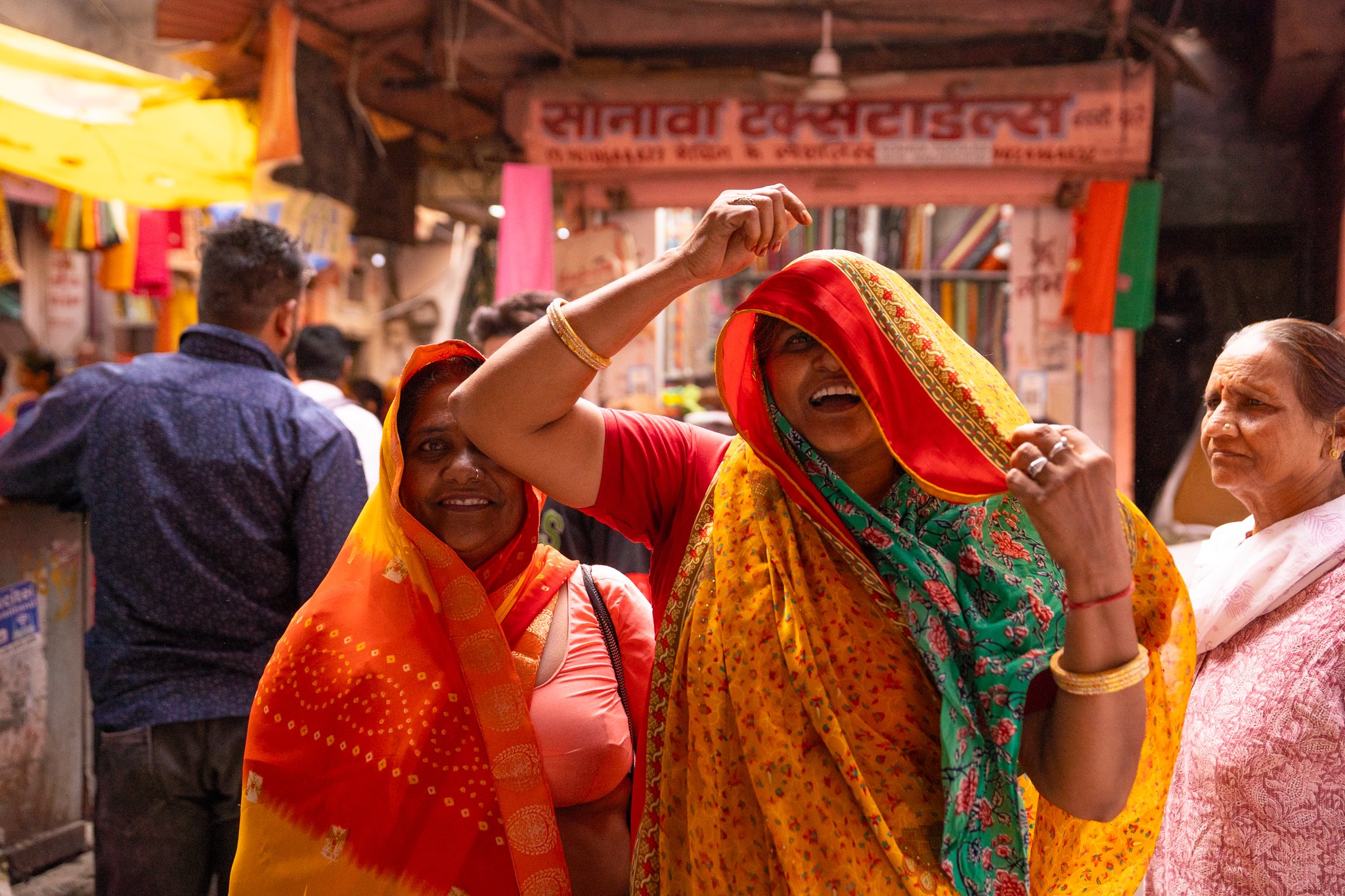 Two women in vivid orange and yellow saris laughing together in a crowded Jaipur market street.