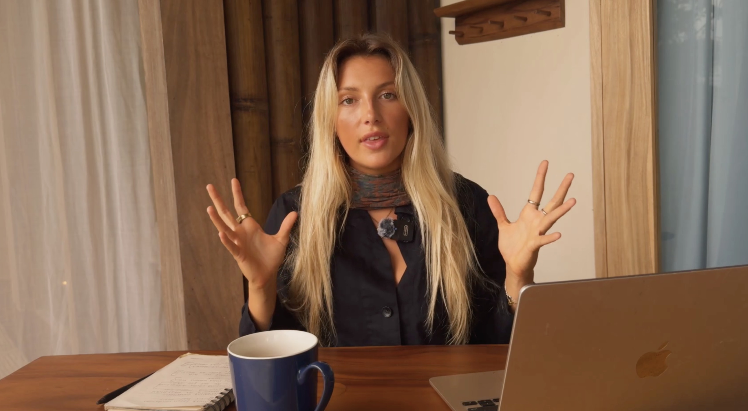 Anna Bucklar sitting at a wooden table with a notebook, mug and laptop, speaking warmly to the camera with both hands open