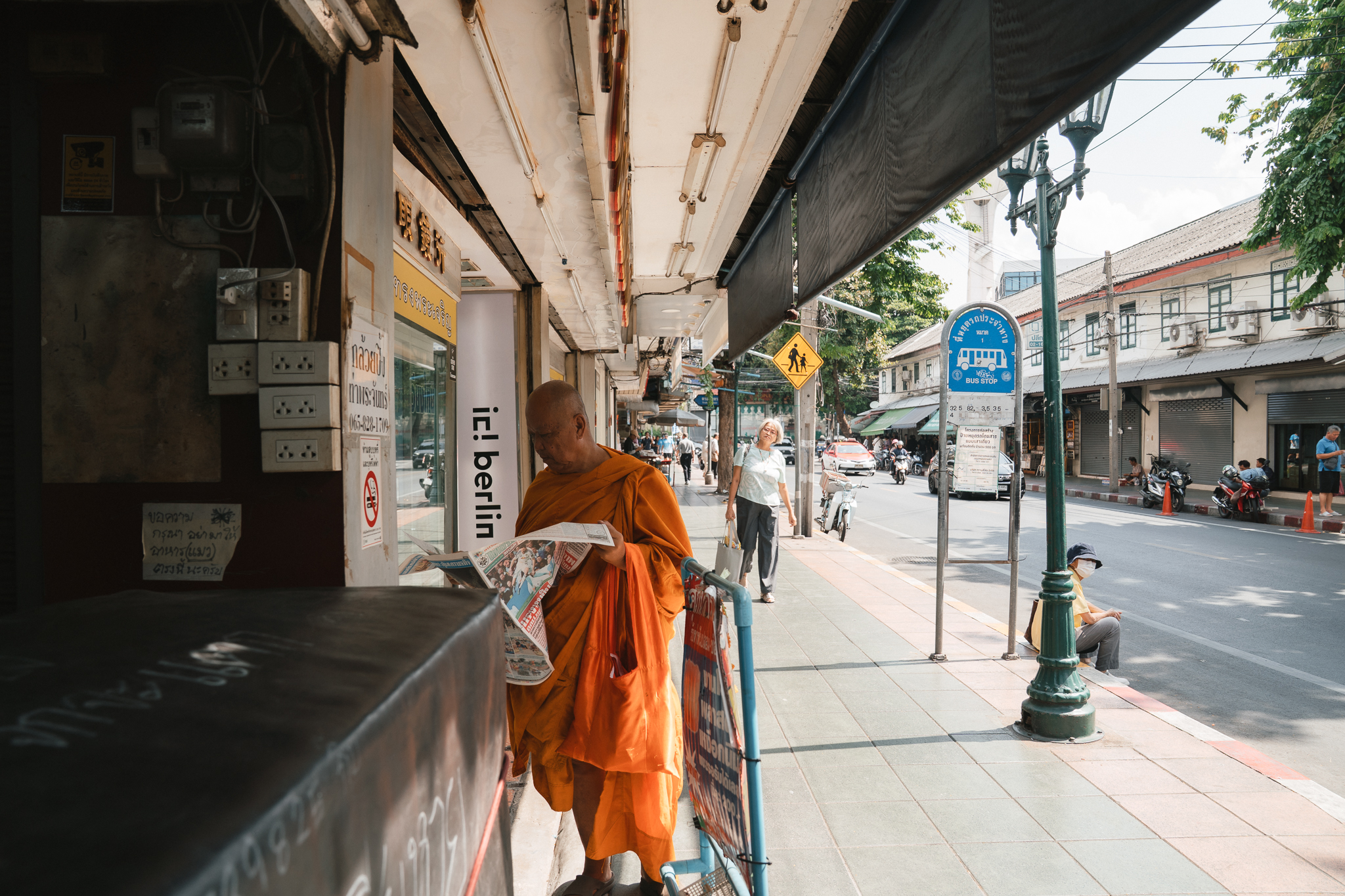 A monk reads the day's news at a quiet bus stop along Tha Phra Chan, the city stirring awake around him.