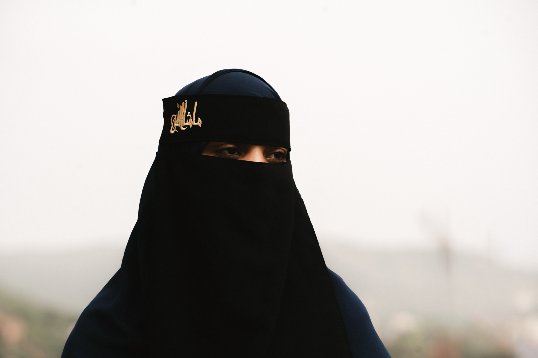 Portrait of a woman in a black niqab with gold embroidery against a soft, hazy sky