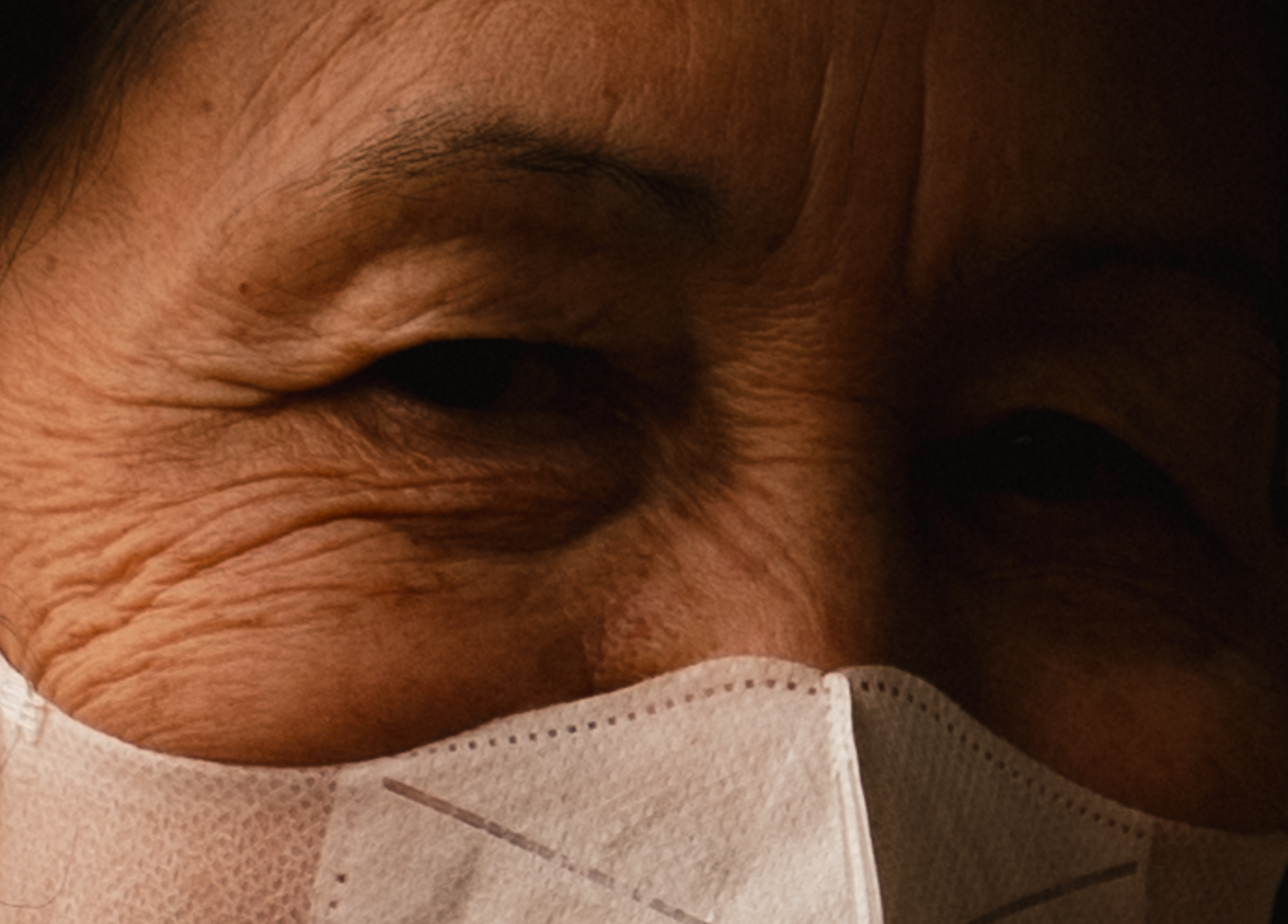 An extreme close-up of an elderly person's weathered, wrinkled eyes above a white mask.