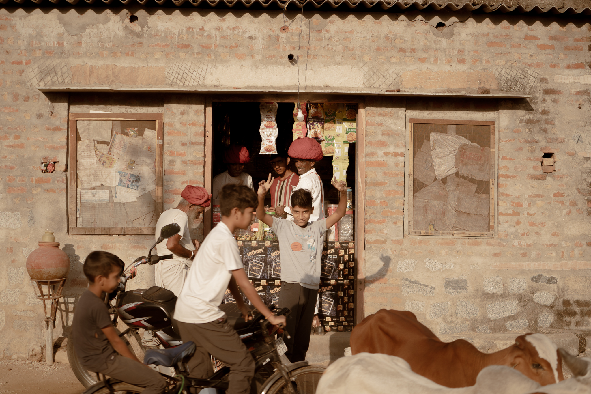Boys mug for the camera outside a village provisions shop while turbaned elders look on and a cow ambles past.